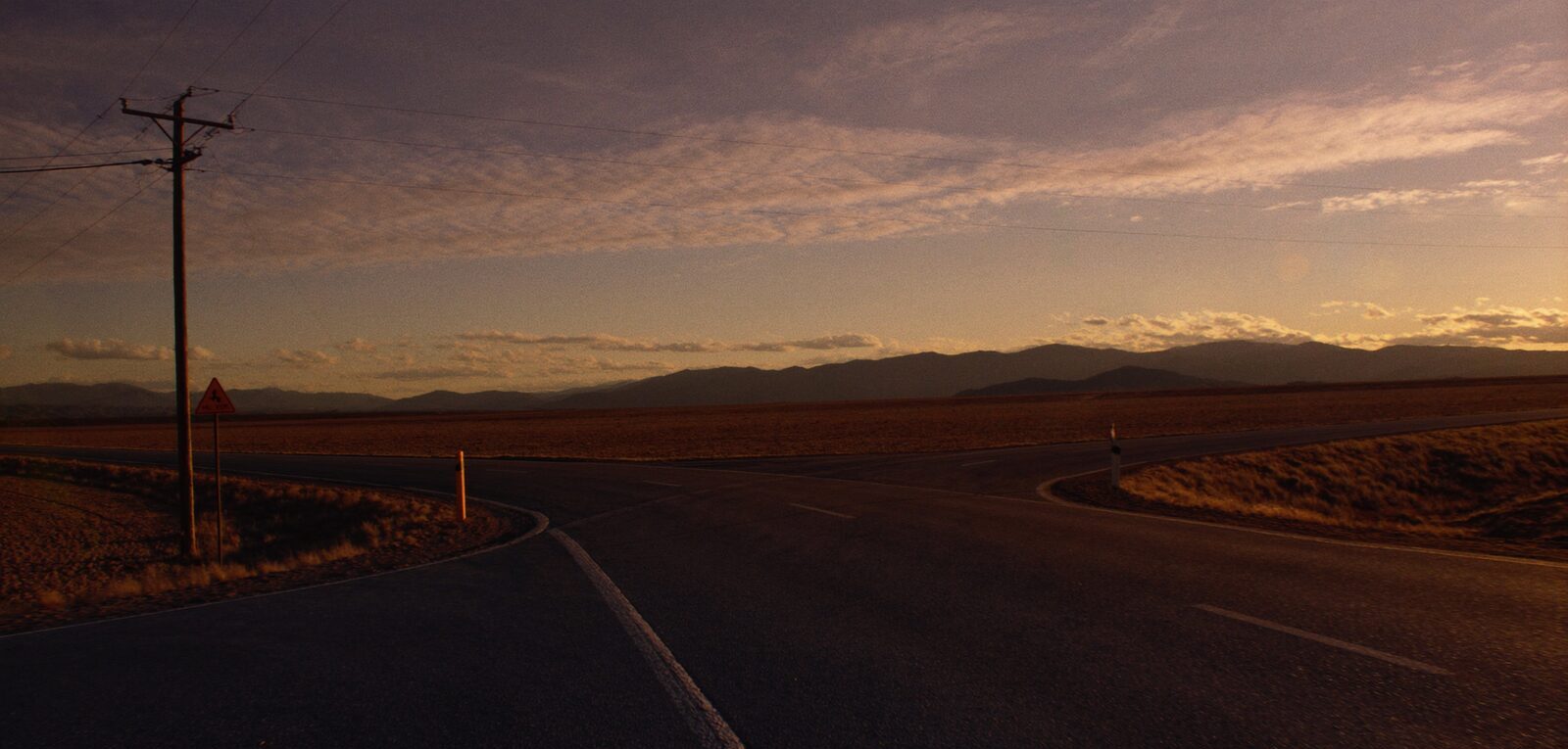 A rural crossroads at golden hour where a road forks toward open plains and distant mountains